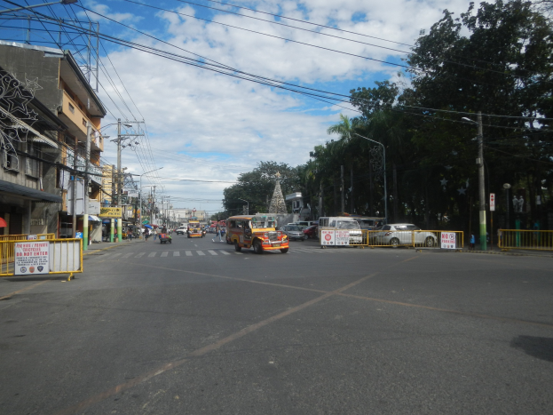 A busy city street with cars, buses, trucks, barricades, signs, light poles, electric poles, buildings, trees, and a cloudy sky.