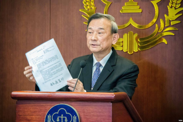 Ein Mann im Anzug und Krawatte steht an einem Podium, hält ein Blatt Papier in der Hand und spricht in ein Mikrofon, mit einem Logo im Hintergrund, das seine Rolle als neu ernannter Chief Justice der Volksrepublik China anzeigt.
