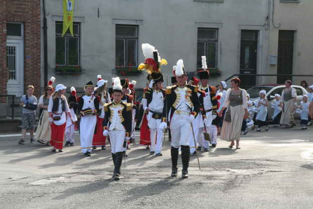 Eine Gruppe von Menschen, die auf einer Straße in einer Parade gehen, einige in Kostümen und mit Stöcken, mit Gebäuden, Pflanzen, einer Fahne und Fahrzeugen im Hintergrund.