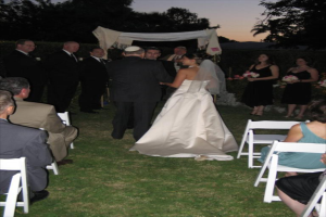 Eine Gruppe von Menschen, die bei einer Hochzeit stehen, wobei die Frauen rechts Blumensträuße halten, während andere auf Stühlen sitzen und Bäume im Hintergrund zu sehen sind.