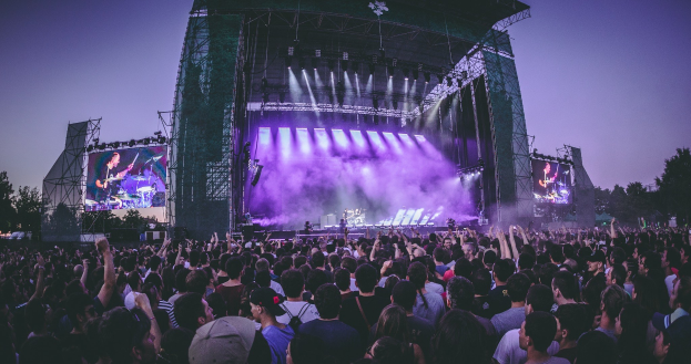 Eine große Menschenmenge bei einem Musikfestival schaut auf einer hell erleuchteten Bühne mit Instrumenten und Bildschirmen, vor einem Gebäude, Bäumen und einem klaren blauen Himmel.