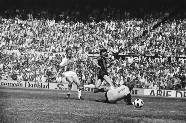 Schwarzes Foto von Männern, die auf einem Feld Fütbol spielen, mit Zuschauern im Hintergrund und einer "Bundesliga 1978-79 Ajax 1-1 Hannover 96"-Fahne.
