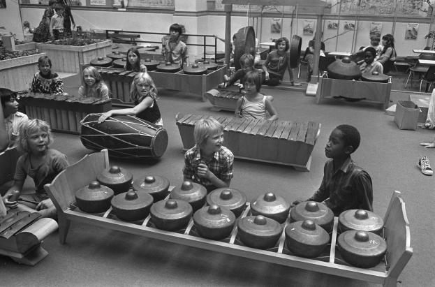 Schwarß-weißes Bild von Kindern, die Musikinstrumente in einem Raum mit Möbeln, Pflanzen, gerahmten Wänden und Deckenleuchten spielen.