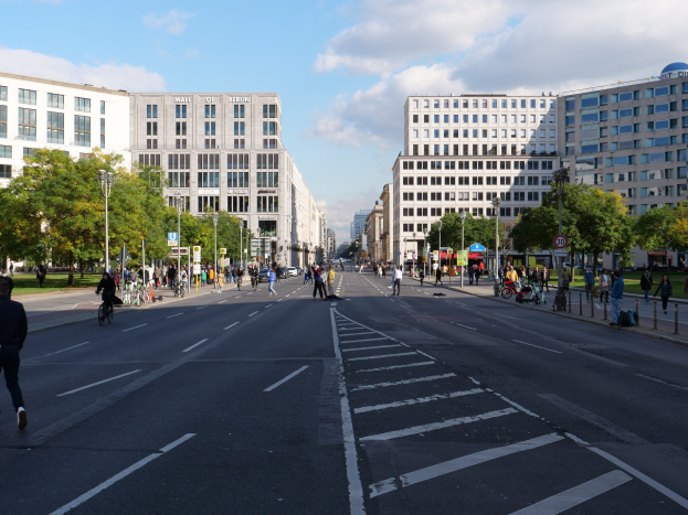 Eine belebte Stadtstraße in Berlin, Deutschland, mit Fußgängern und Radfahrern auf der Straße, hohen Gebäuden mit Fenstern, Bäumen, Laternen und Schildern.