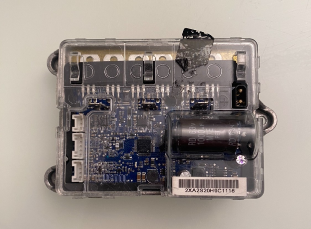 Close-up of a small electronic device in a clear plastic case on a white surface, showing a circuit board with a battery attached.