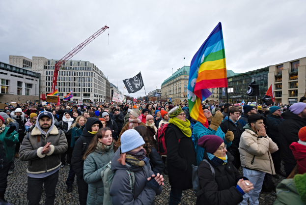 Eine große Gruppe von Menschen mit Fahnen und Schildern mit Text, darunter "Lgbtq+ Rechte Demonstration in Berlin", die vor einem Gebäude mit einem Kran und einem bewölkten Himmel stehen.