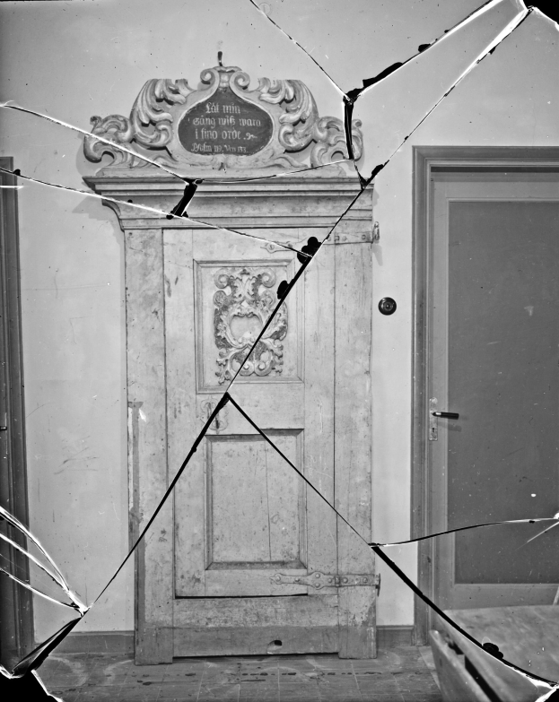 Schwarz-weiß-Foto einer kaputten Tür in einem Raum mit einer Wand im Hintergrund, die Tür in einem desolaten Zustand mit verstreuten Glasscherben.