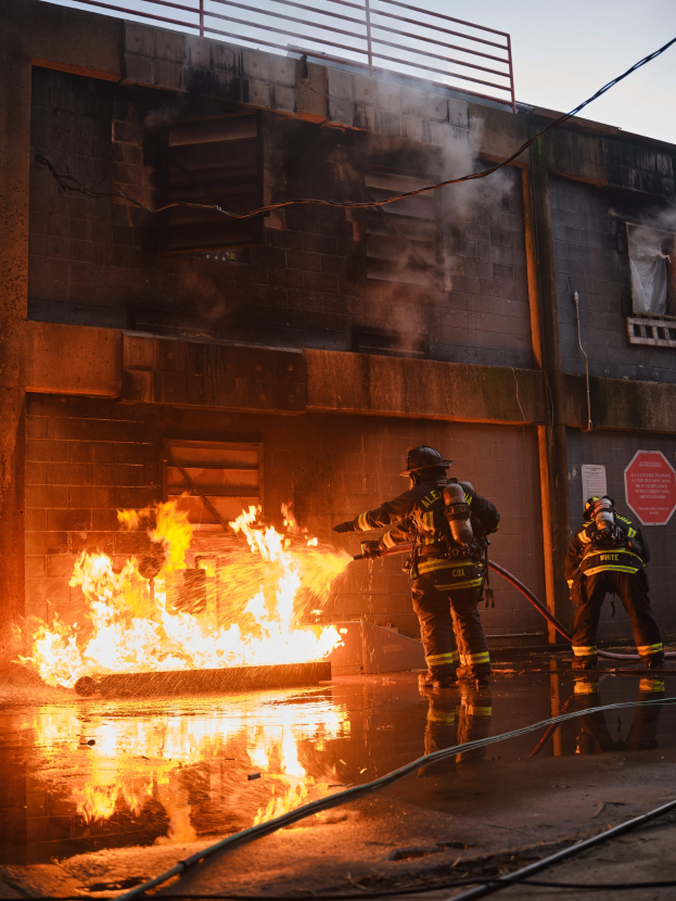 Drei Feuerwehrleute in Helmen arbeiten daran, ein Feuer in einem brennenden Gebäude zu löschen, mit Rauch, der aus dem Dach entweicht und einem Schild an der Wand.