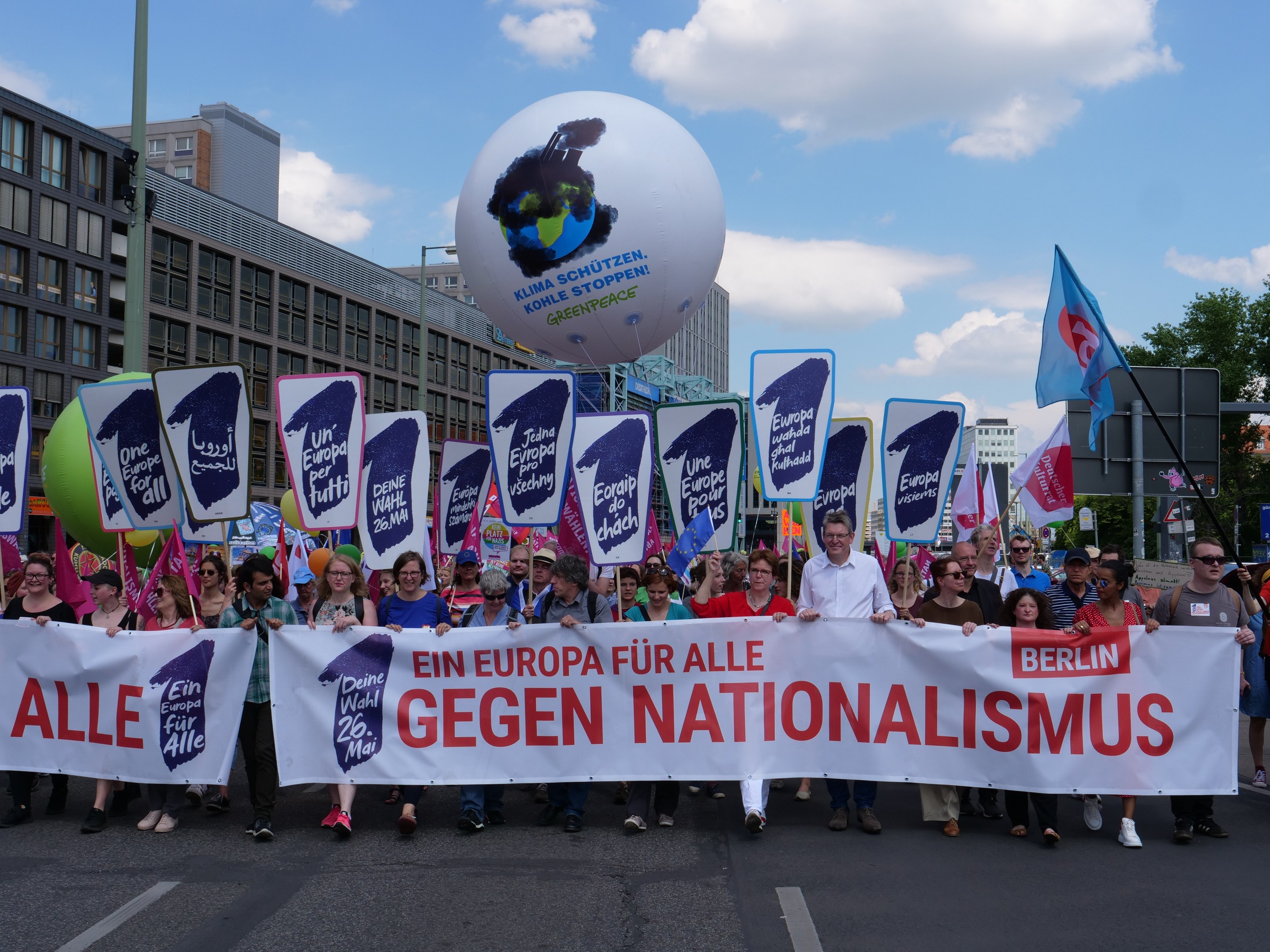 Group of people walking down a street holding a banner reading "Ein Europa für alle Gegen Nationalismus" and flags, with buildings, trees, and a cloudy sky in the background.