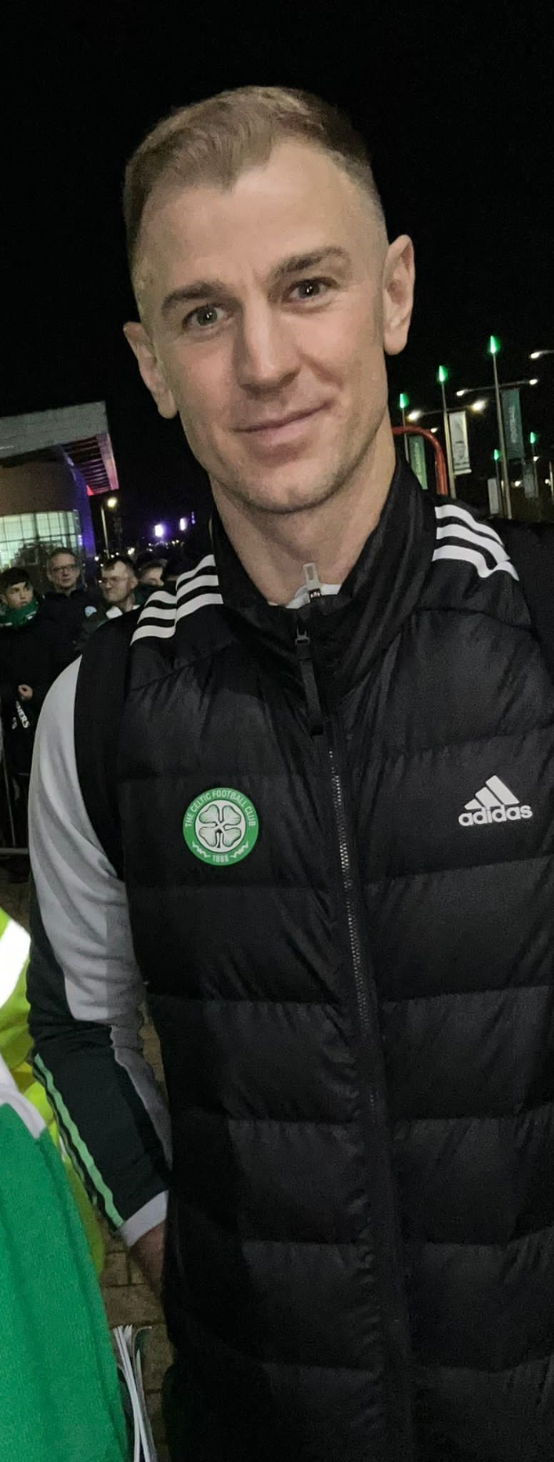Ein Mann in einer schwarzen Jacke steht lachend vor einer Menge, beleuchtet von Gebäudelichtern und Himmel, wahrscheinlich feiernd nach Celtic's Sieg im Scottish Cup Final.