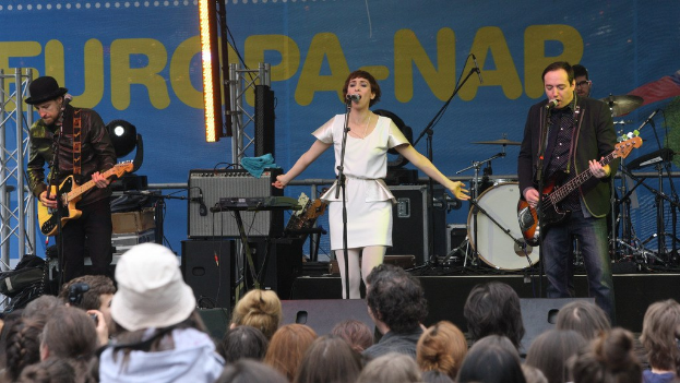 Vier Personen auf der Bühne: eine Frau in einem weißen Kleid singt in der Mitte, flankiert von zwei Männern mit Gitarren, mit einer Menge unten und Lautsprechern gegen eine Wand im Hintergrund.