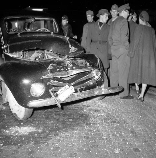 Schwarz-weißes Bild eines schwer beschädigten Autos am Straßenrand mit einer Gruppe von Menschen, die es inspizieren.