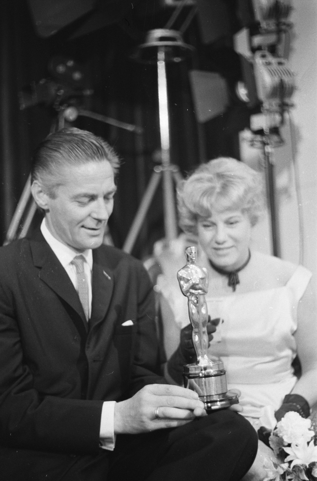 Ein Mann in einem schwarzen Blazer, weißem Hemd und Krawatte hält eine Oscar-Statue in der Hand und sitzt neben einer Frau, die einen Blumenstrauß hält, beide in eleganter Kleidung, mit abgedunkeltem Hintergrund.