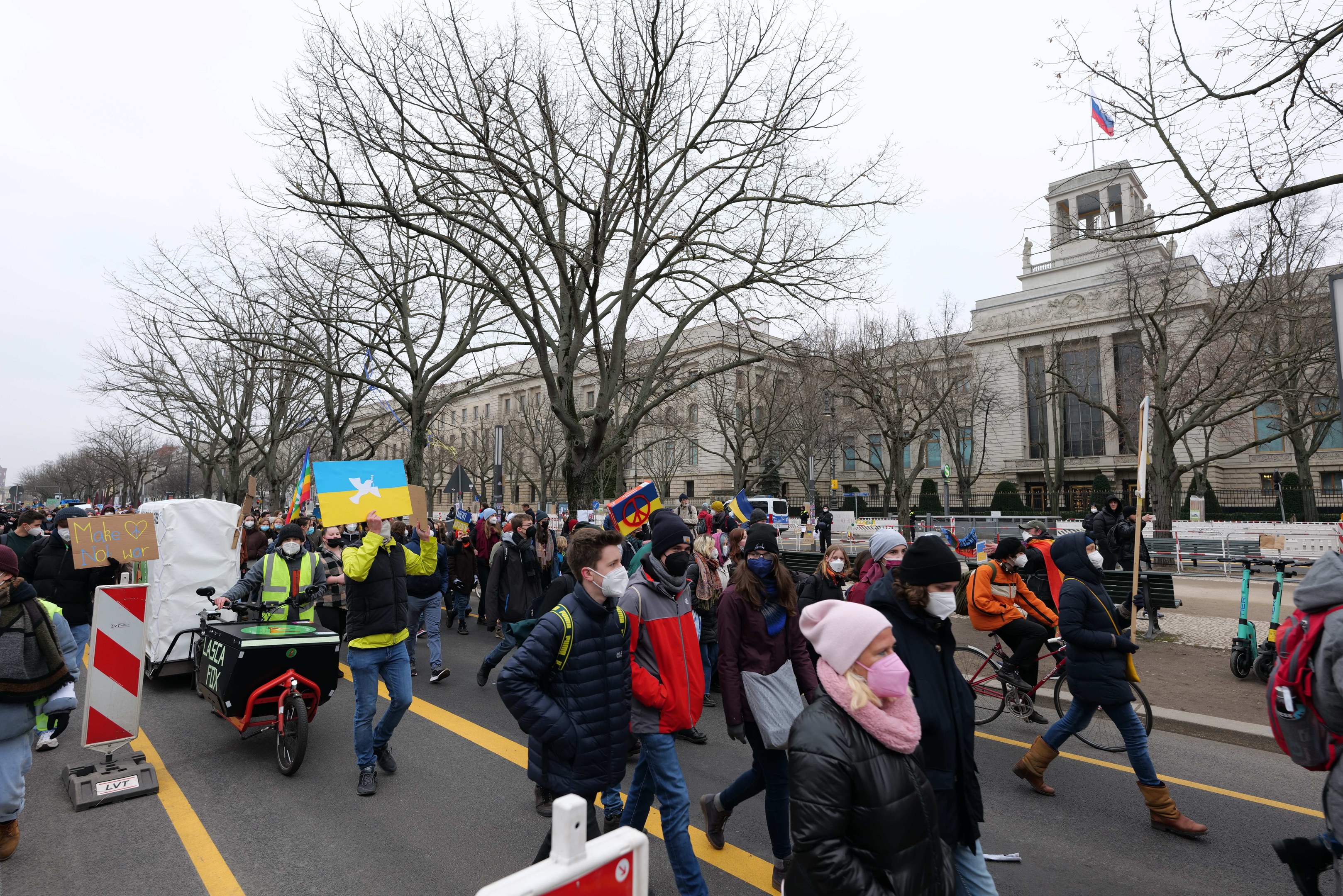 A large group of people marching in a protest on a city street, some holding signs and others riding bicycles, with trees and a building in the background under a clear blue sky.