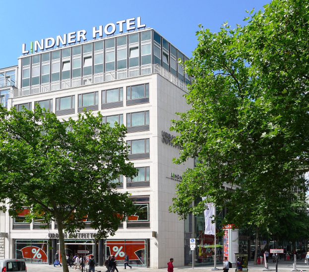 Das Lindner Hotel in Berlin, ein großes Glasfrontgebäude mit Beschriftung oben, umgeben von Bäumen, Fahrzeugen und Fußgängern unter einem sichtbaren Himmel.