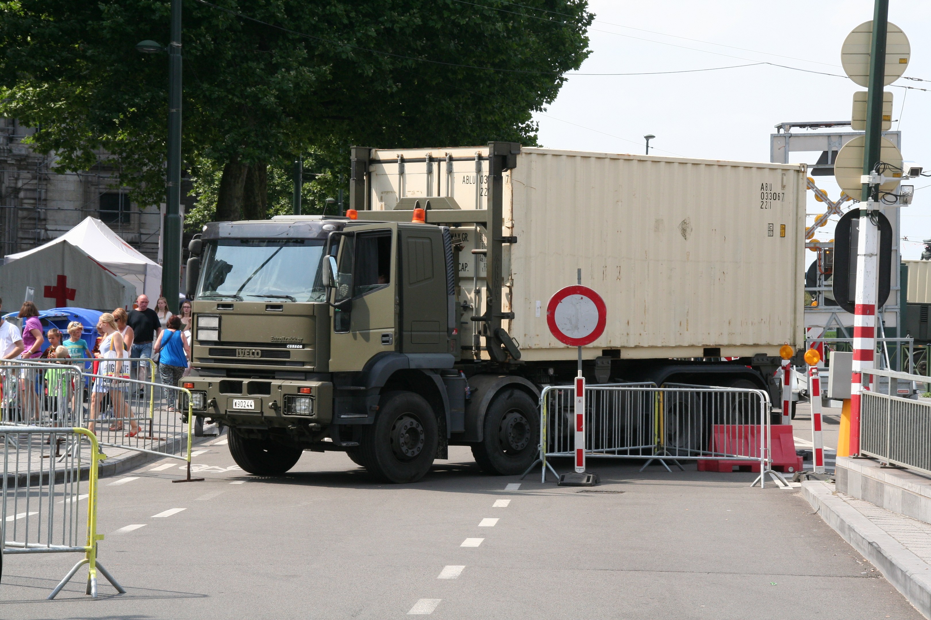 Ein Lkw am Straßenrand mit einer Gruppe von Menschen auf dem Gehweg, umgeben von Absperrungen, Schildern, Straßenlaternen, Strommasten, Stromkabeln, Bäumen, Gebäuden und einem klaren blauen Himmel.