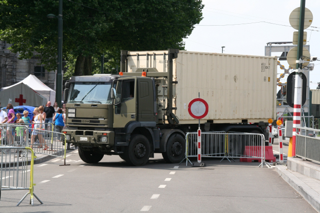 Ein Lkw am Straßenrand mit einer Gruppe von Menschen auf dem Gehweg, umgeben von Absperrungen, Schildern, Straßenlaternen, Strommasten, Stromkabeln, Bäumen, Gebäuden und einem klaren blauen Himmel.