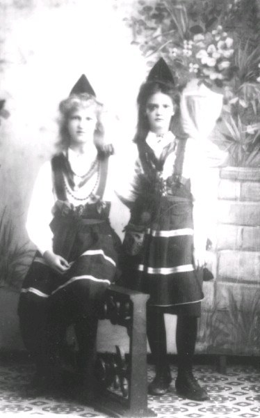 Two young girls in traditional Bavarian clothing sit on a bench with a plant-covered wall in the background, captured in black and white.