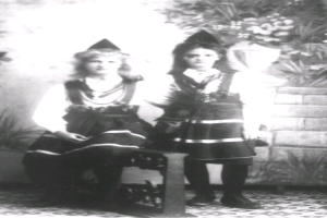 Two young girls in traditional Bavarian clothing sit on a bench with a plant-covered wall in the background, captured in black and white.