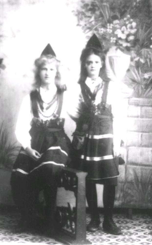 Two young girls in traditional Bavarian clothing sit on a bench with a plant-covered wall in the background, captured in black and white.
