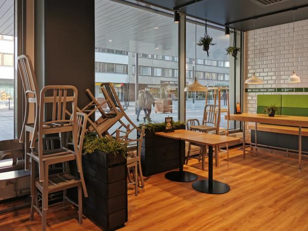 Ein Berliner Restaurant mit Holztischen und -stühlen in der Nähe eines großen Fensters, das städtische Elemente wie Fußgänger, Gebäude und Straßenmöbel zeigt.