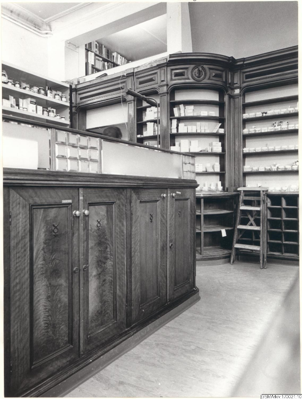 Schwarzes und wei├čes Foto eines vintage Apothekeninneren mit einer Theke, Sch├Ąben, einer Leiter, Regalen mit Beh├Ąltern und Text überall.