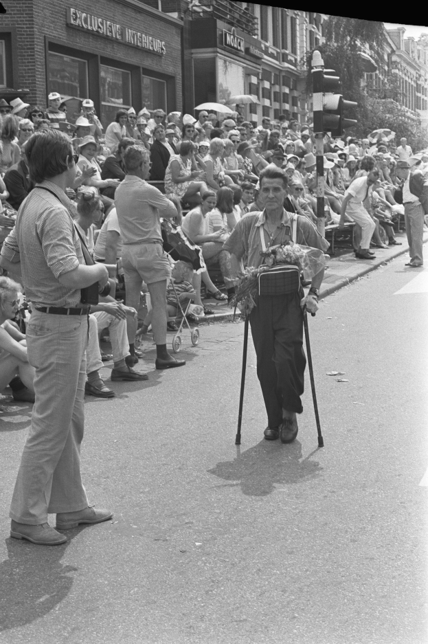 Schwarzes und weißes Bild eines Mannes, der mit einem Stock die Straße entlanggeht, vor einer Menge von Menschen, einige sitzen und einige stehen, mit einer Ampel, Gebäuden und Bäumen im Hintergrund.
