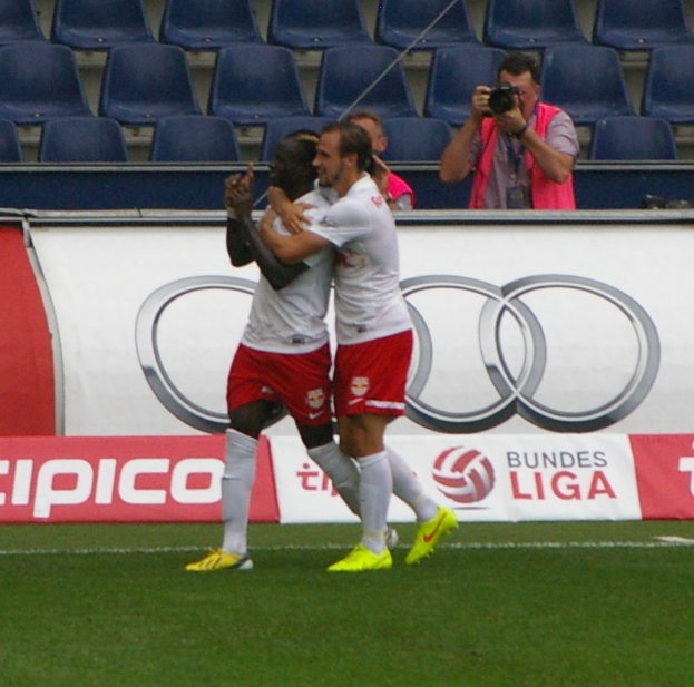 Zwei Fussballspieler feiern ein Tor auf einem Feld mit Zuschauern, einem Fotografen, einem Banner, Stühlen und einer Wand im Hintergrund.