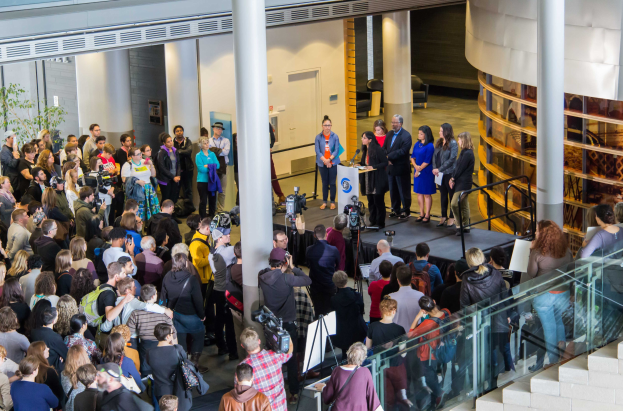 Eine Gruppe von Menschen steht auf einer Treppe, die zu einem Podium mit einem Mikrofon führt, umgeben von einer Menge mit Kameras auf der 2018 Nationalen Frauen-Gesundheitskonferenz.