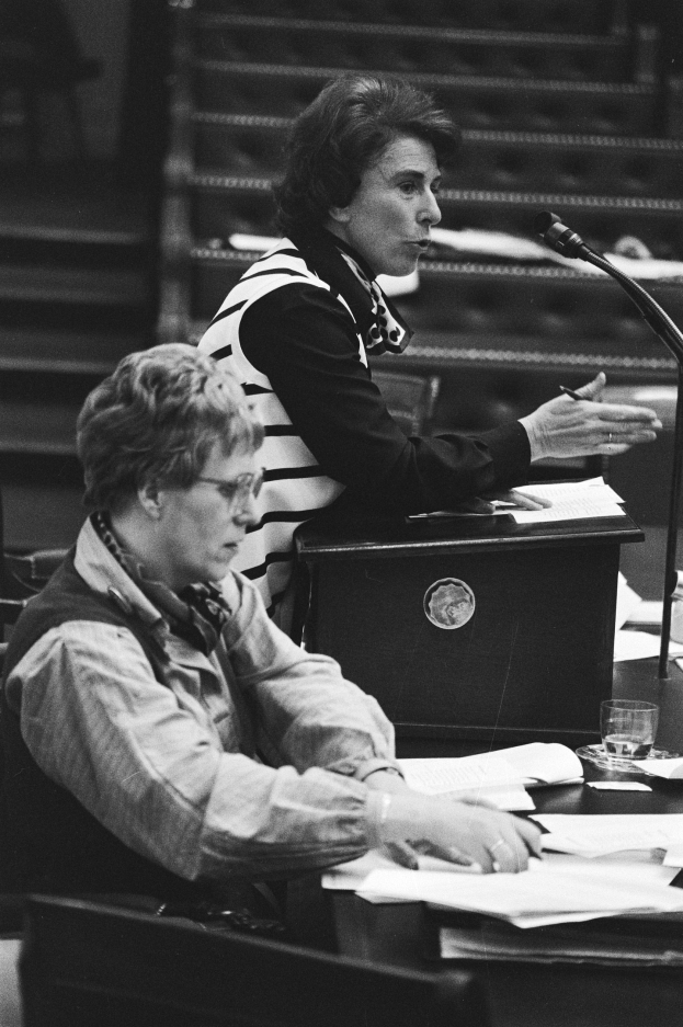 Schwarz-weißes Foto einer Frau, die an einem Tisch im Europäischen Parlament in Straßburg sitzt und einen Stift hält, mit Papieren, einem Glas Wasser und anderen Gegenständen auf dem Tisch, Treppen im Hintergrund.