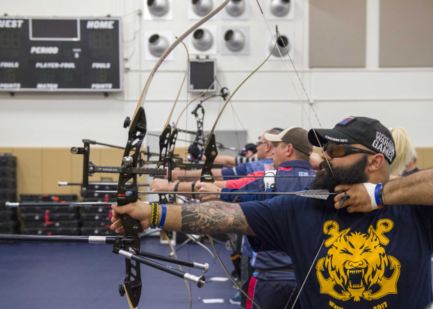 Eine Gruppe von Bogenschützen in Mützen übt drinnen während der World Archery Championships 2017, mit einem Textboard links und Equipment rechts.
