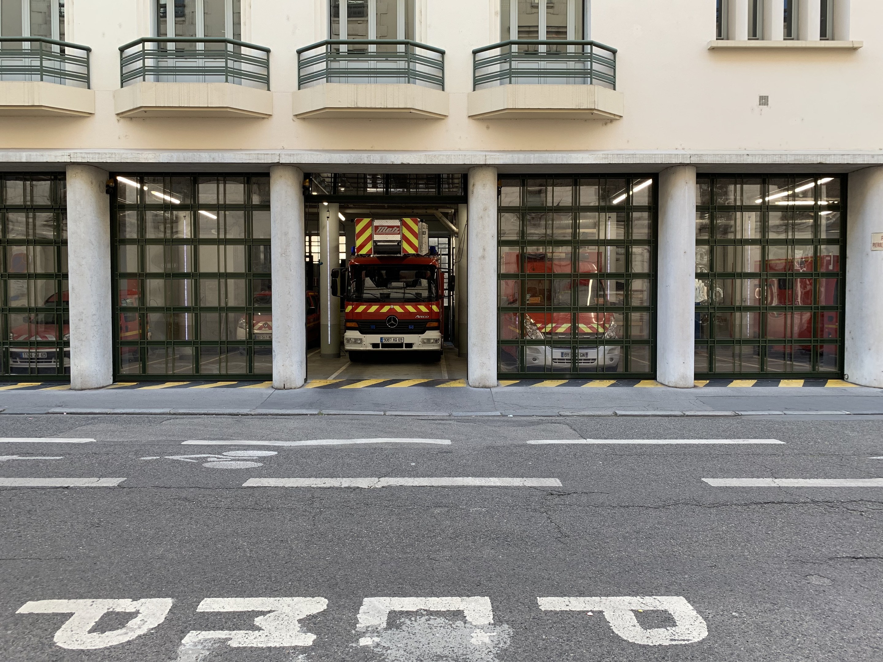 Feuerwache mit Feuerwehrauto davor, mit Fenstern, Geländern, Säulen, Glastüren und einer Straße unten.