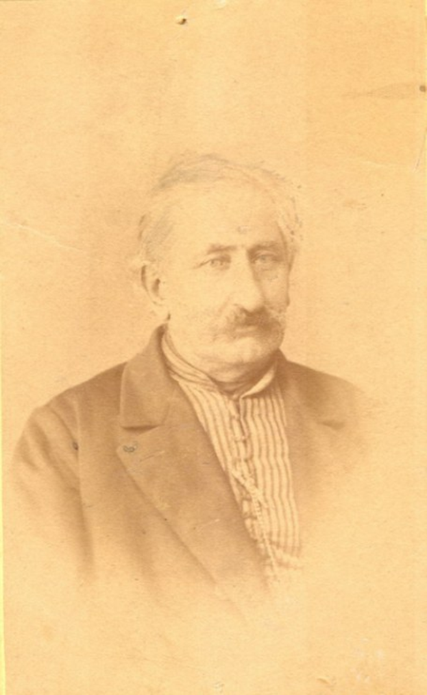 Ein Schwarz-Weiß-Porträt eines älteren Mannes mit Schnurrbart, der einen Mantel trägt und ein starkes Kinn, eine markante Nase und einen nachdenklichen Ausdruck mit leicht geöffneten Lippen hat.