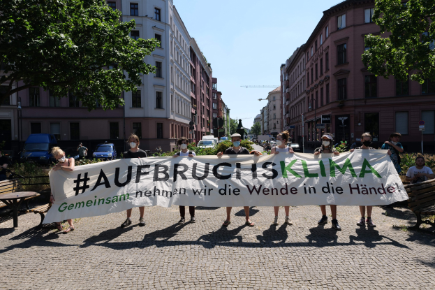A group of people wearing masks holding a banner reading "Aufbruchsklima" during a climate change protest in Berlin, Germany, with surrounding urban elements visible.