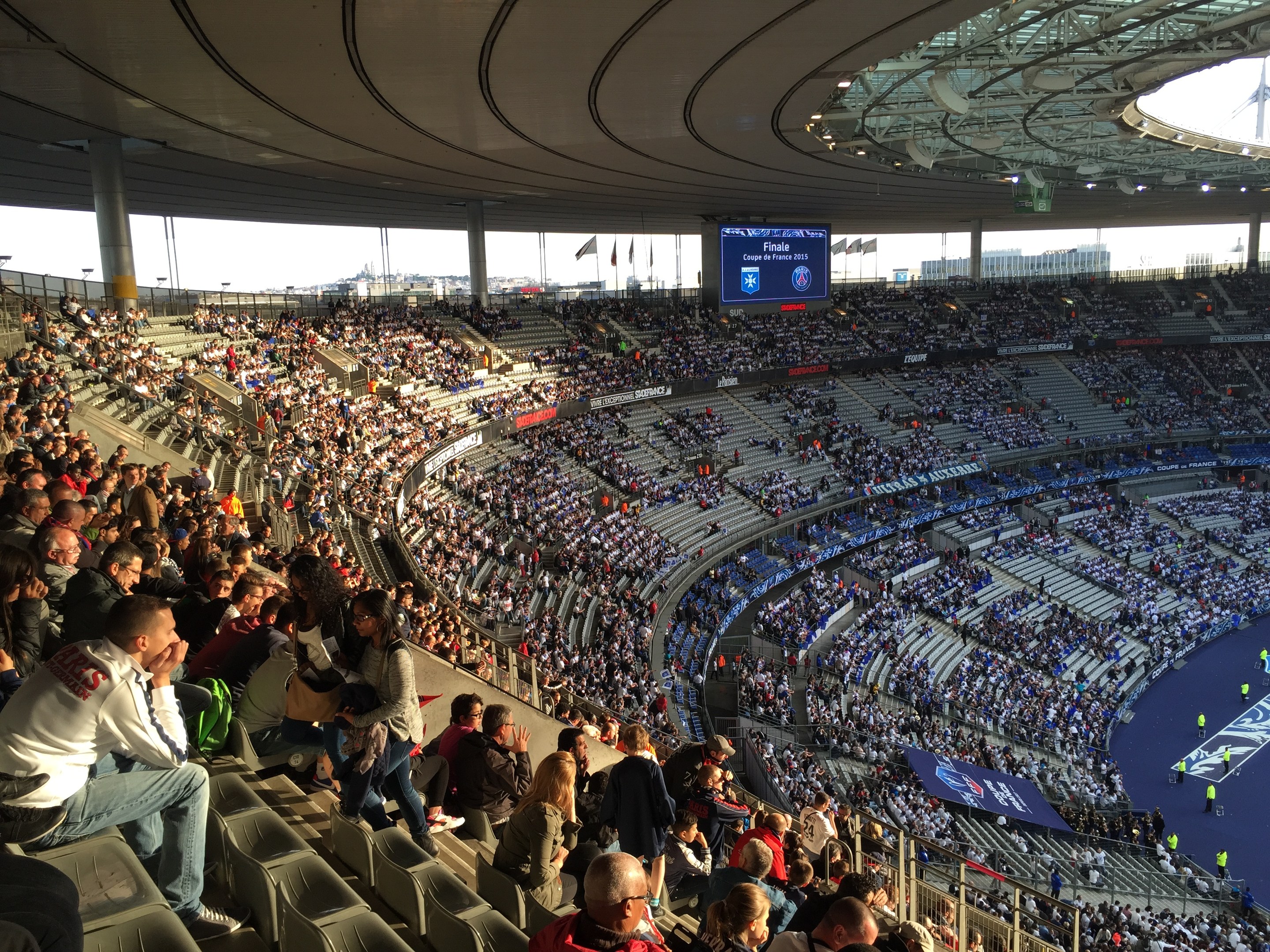 Große Menschenmenge in einem Stadion bei einem Fußballspiel mit einer Bühne, Fahnen, Stangen, einem Bildschirm und der Allianz Arena in München, Deutschland im Hintergrund.