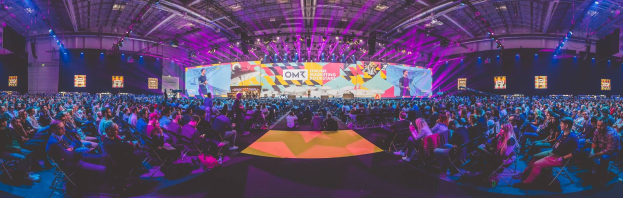 Eine große Zuschauermenge sitzt auf Stühlen einem hell erleuchteten Bühne mit Bildschirmen gegenüber, auf der mehrere Menschen stehen während eines Online-Marketing-Events.