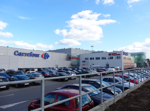 Großer Autohandel mit zahlreichen parkenden Fahrzeugen, umgeben von städtischen Gebäuden, Straßeninfrastruktur und Fahrzeugen auf der Straße unter einem bewölkten Himmel.