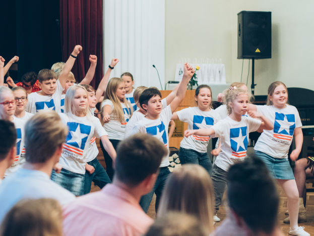 Eine Gruppe von Kindern in weißen Hemden und blauen Shorts tanzt vor einem sitzenden Publikum, mit einer Vorhangwand, einem Lautsprecherständer und einem Tisch mit Mikrofon im Hintergrund.
