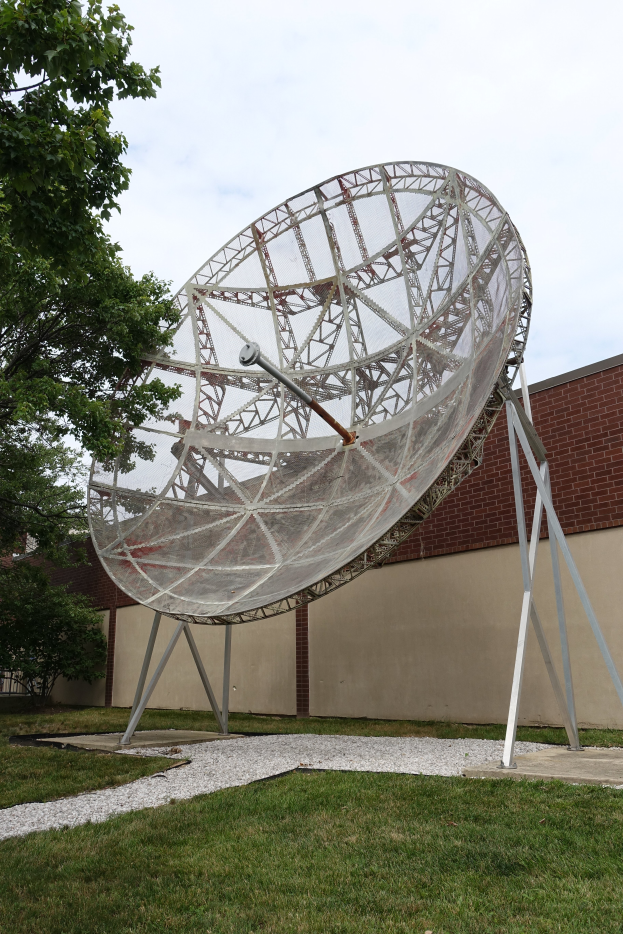 Eine große Satellitenschüssel auf einem Rasenfeld mit einem Baum links und einem Gebäude im Hintergrund unter einem sichtbaren Himmel.