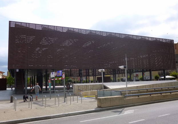 Großes modernes Gebäude mit zahlreichen Fenstern als neuer Eingang zu einer Stadtmitte, umgeben von städtischer Infrastruktur einschließlich Straßenlaternen, Fahrzeugen, Fußgängern und Bäumen unter einem bewölkten Himmel.