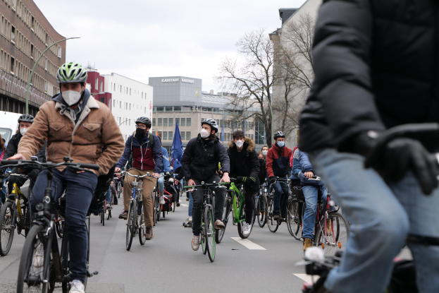 Eine Gruppe von Menschen in Helmen und Handschuhen fährt mit Fahrrädern eine von Bäumen gesäumte Straße in Berlin, Deutschland, entlang, wobei Gebäude und ein geparktes Fahrzeug zu sehen sind.