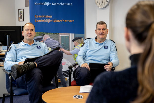 Two police officers seated at a table with a woman, surrounded by papers and objects, with a wall-mounted banner, clock, and photo frames in the background, and another table with a monitor and accessories to the left.