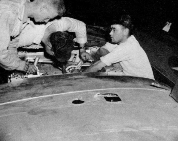 Drei Männer in Kappen reparieren ein schwarz-weißes Auto in einer Garage, umgeben von Werkzeugen und Materialien.