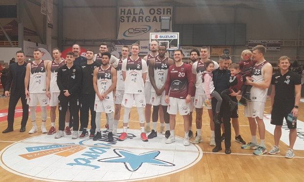Mannergruppe auf einem Basketballfeld mit Kindern, Zuschauern im Hintergrund, Bannern mit "Hala Osir Starga", einem Basketballkorb, Metallstangen, Deckenleuchten und einer Wand.