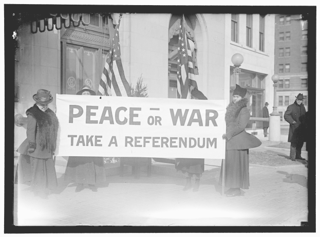 Gruppe von Frauen mit einem Banner mit der Aufschrift "Frieden oder Krieg, machen Sie eine Volksabstimmung" vor einem Gebäude mit Fenstern, einer Pflanze, einem Straßenpfahl und einem bewölkten Himmel.