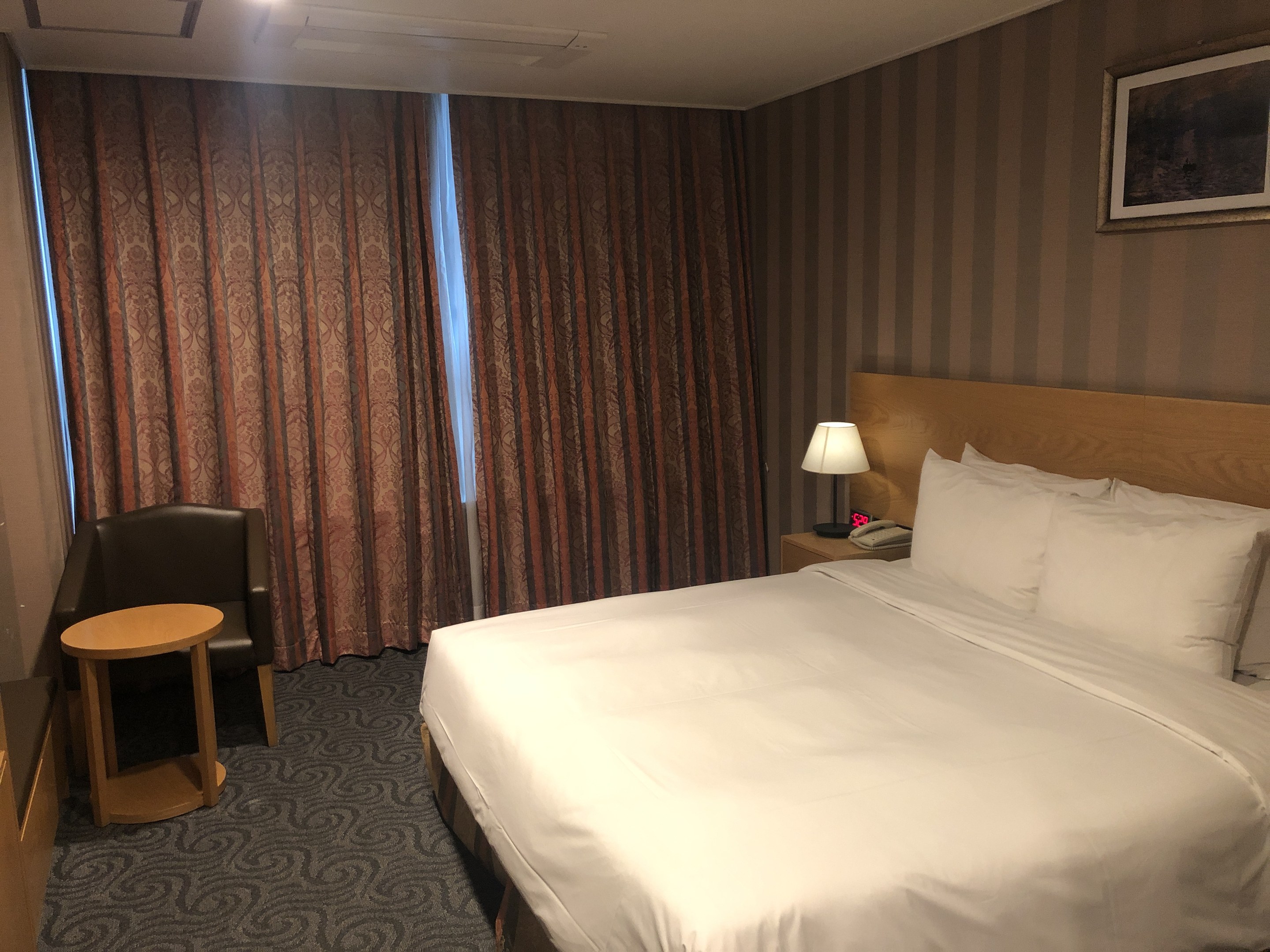 Hotelzimmer mit großem Bett, Stuhl, Tisch, Telefon, Lampe, Foto auf dem Wand, Fenster mit Vorhängen und Deckenleuchten.