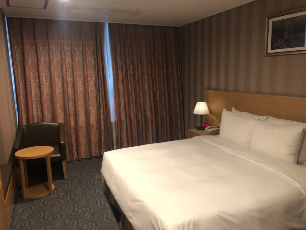 Hotelzimmer mit großem Bett, Stuhl, Tisch, Telefon, Lampe, Foto auf dem Wand, Fenster mit Vorhängen und Deckenleuchten.