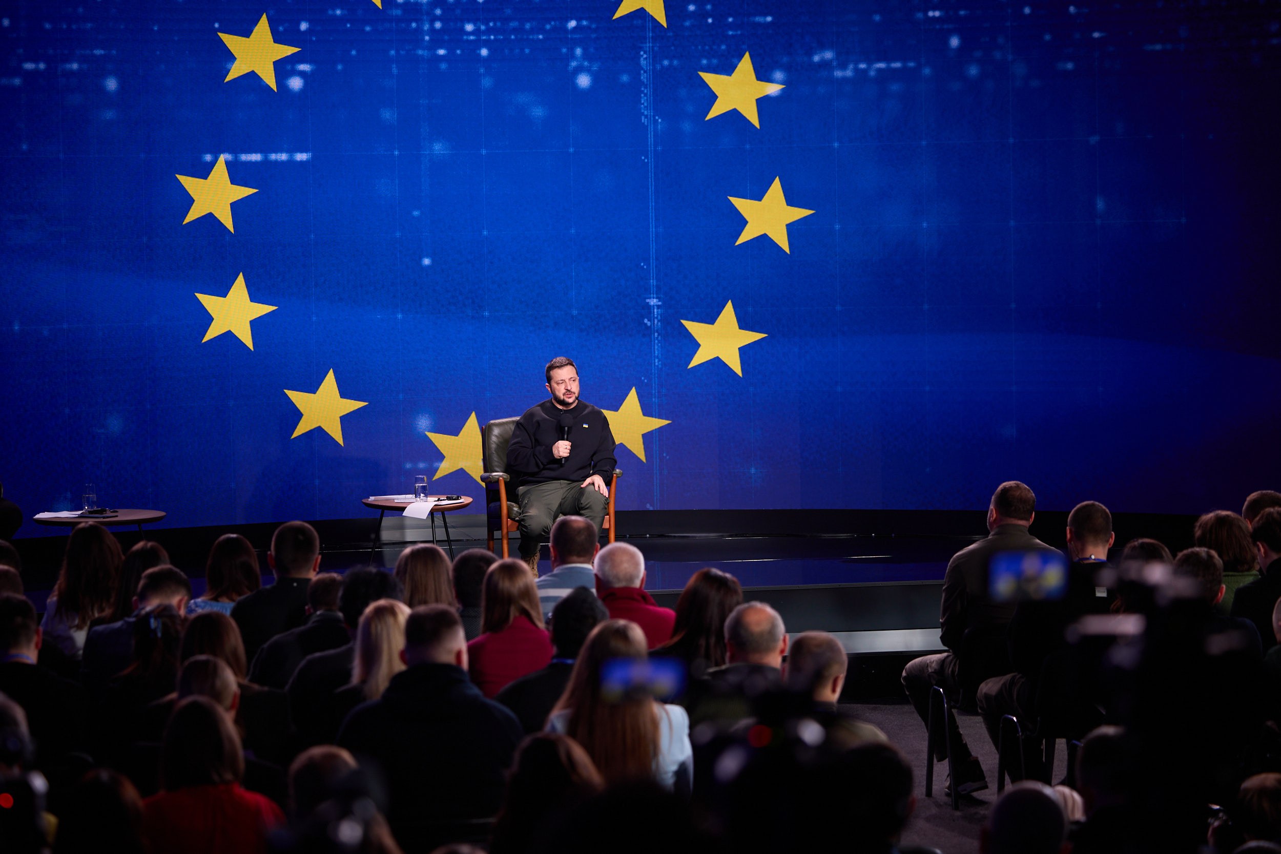 A man in a black dress sits on a stage holding a microphone, speaking to a seated audience, with two tables and a blue screen displaying yellow stars in the background.