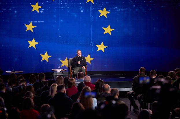 A man in a black dress sits on a stage holding a microphone, speaking to a seated audience, with two tables and a blue screen displaying yellow stars in the background.
