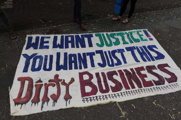 Banner auf einer Straße mit dem Text "Wir wollen Gerechtigkeit, du willst nur diesen schmutzigen Deal" und zwei Personen oben drauf, eine hält ein blaues Objekt.
